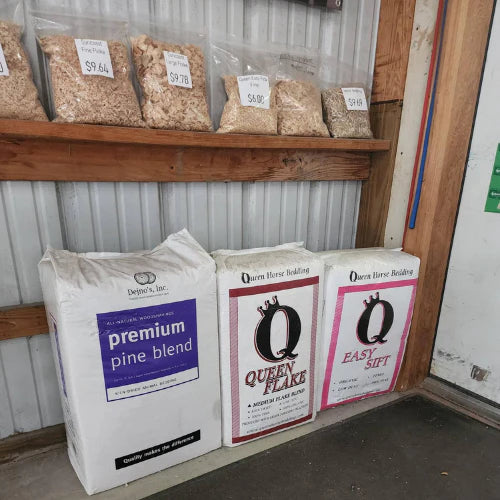 Bags of horse bedding and pine blend are displayed on a floor under a shelf with smaller bags of wood shavings labeled with prices. Industrial setting.